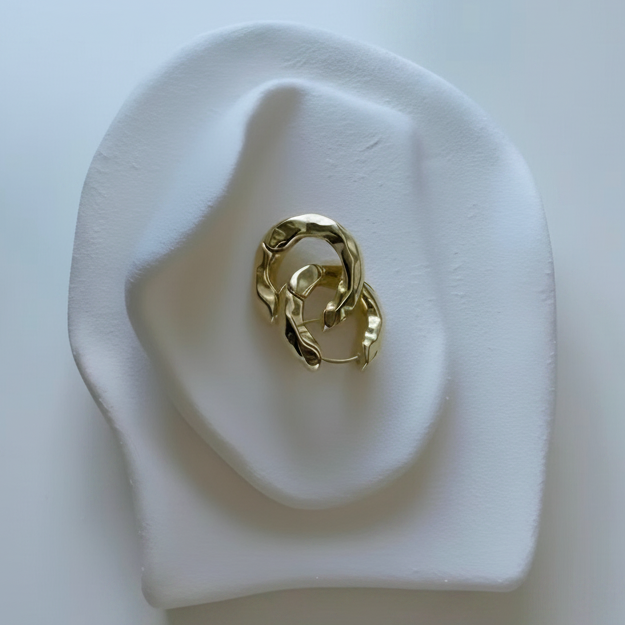 A gold-toned abstract ring with a twisted, organic knot design, displayed on a white sculptural stand against a clean white background.