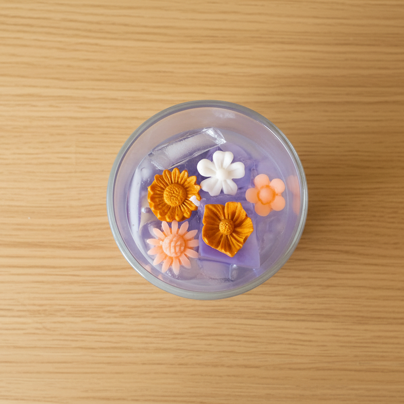A top-down view of a clear round cup containing a translucent purple gel or jelly with three small decorative flower charms in orange, pink, and white, placed on a light wood-grain surface.