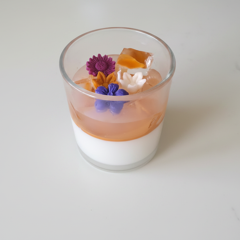 A clear glass cup containing a layered dessert or gel, with a white lower layer, a peachy-orange upper layer, and small edible flowers on top, photographed on a clean white surface.